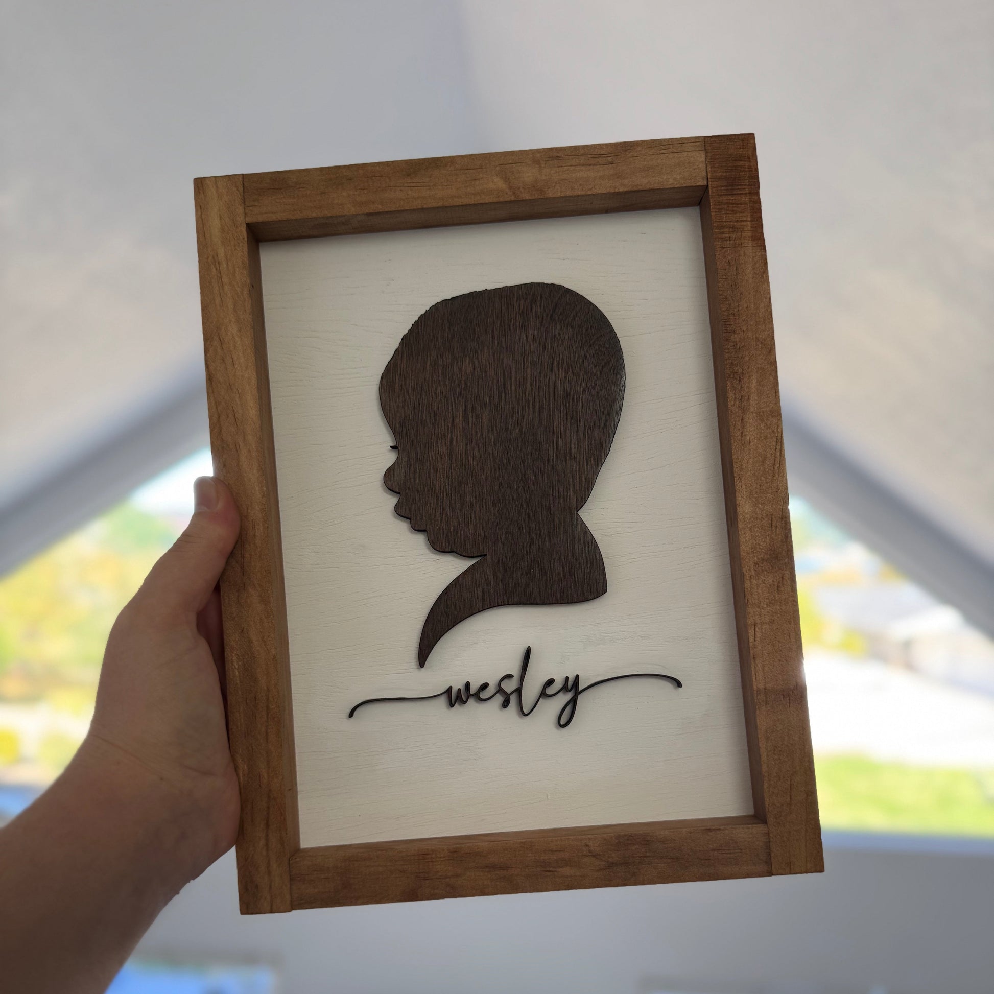 Wooden frame with silhouette and 'Wesley' text held by a hand indoors.