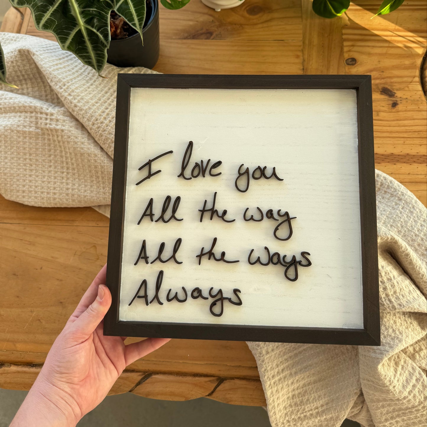 Framed quote 'I love you All the way All the ways Always' held by a hand on a wooden surface with plants in the background.